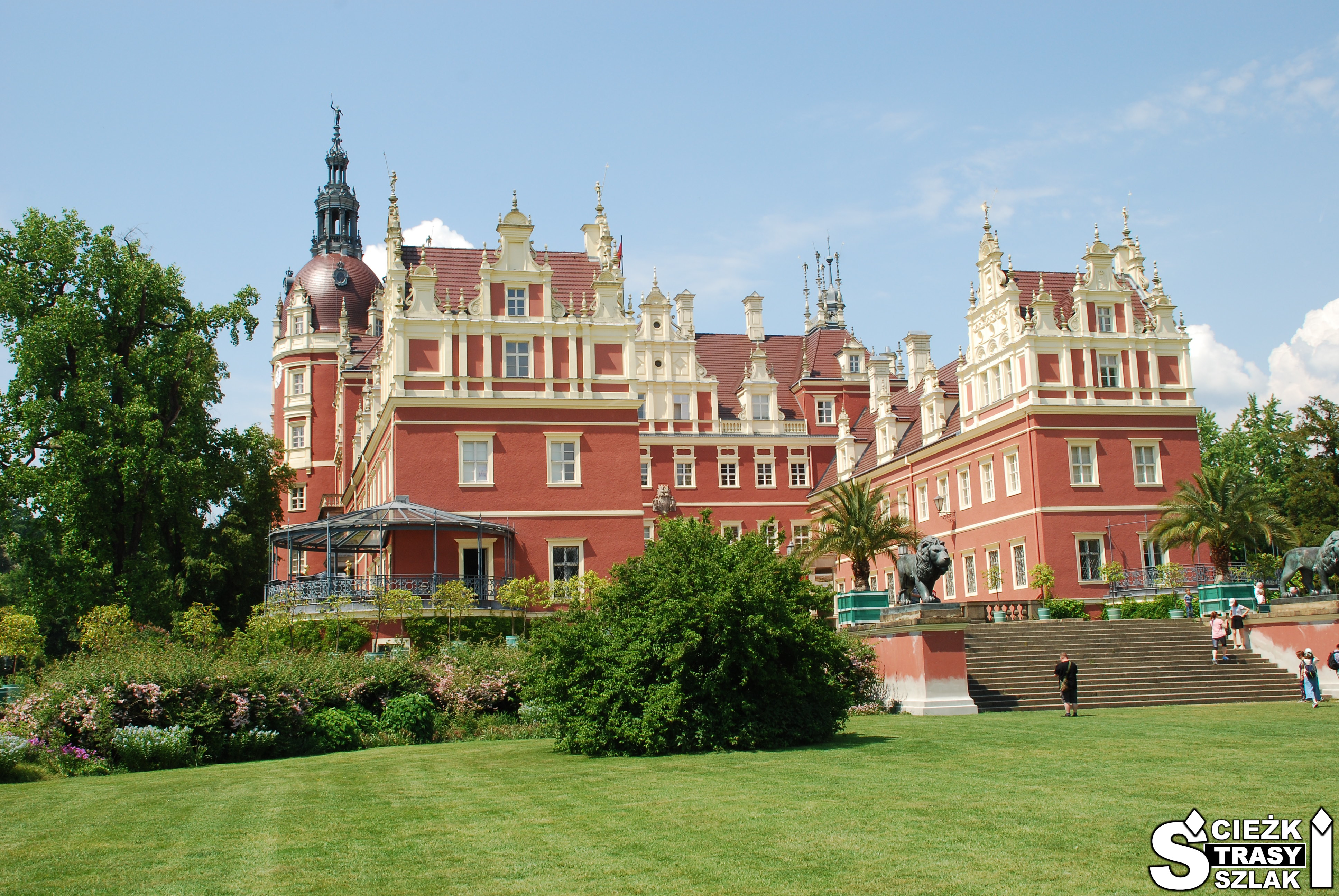 Zamek z wieżami i czerwoną fasadą z wysokimi schodami do zabytkowej budowli wśród drzew i kwitnących krzewów Parku Mużakowskiego