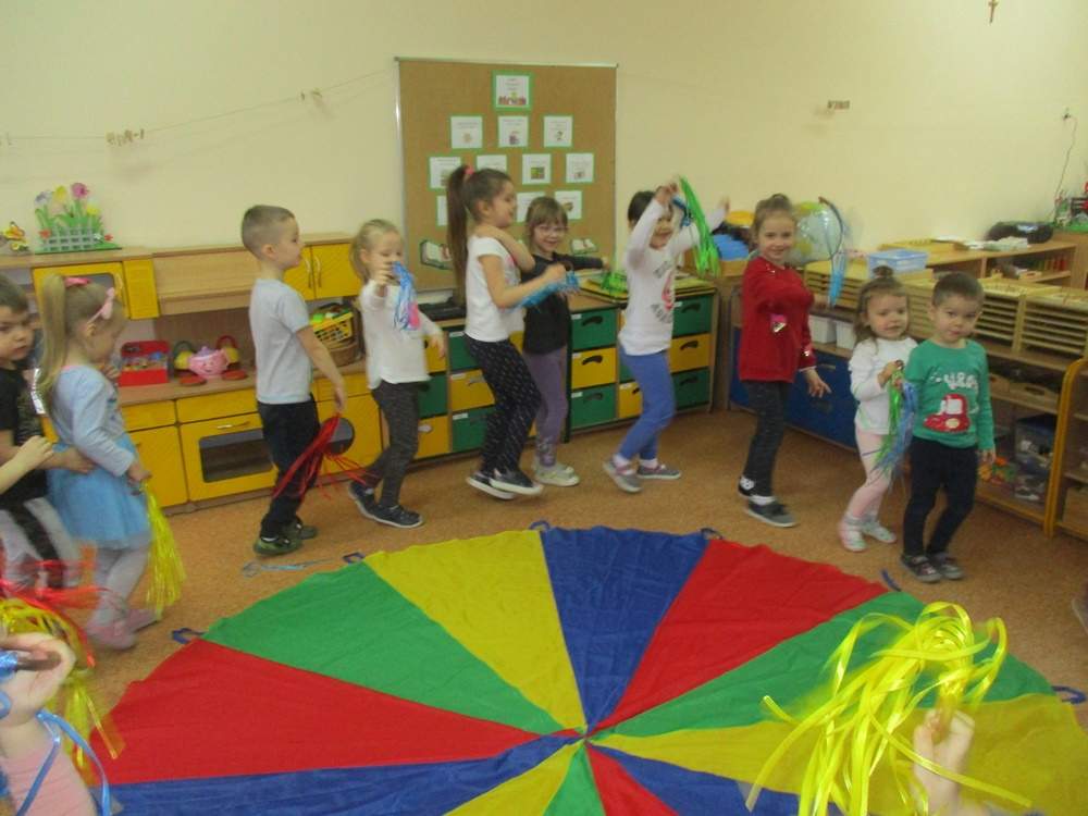 Dzieci  poruszają się wokół chusty w rytm muzyki, machają kolorowymi wstążkami.