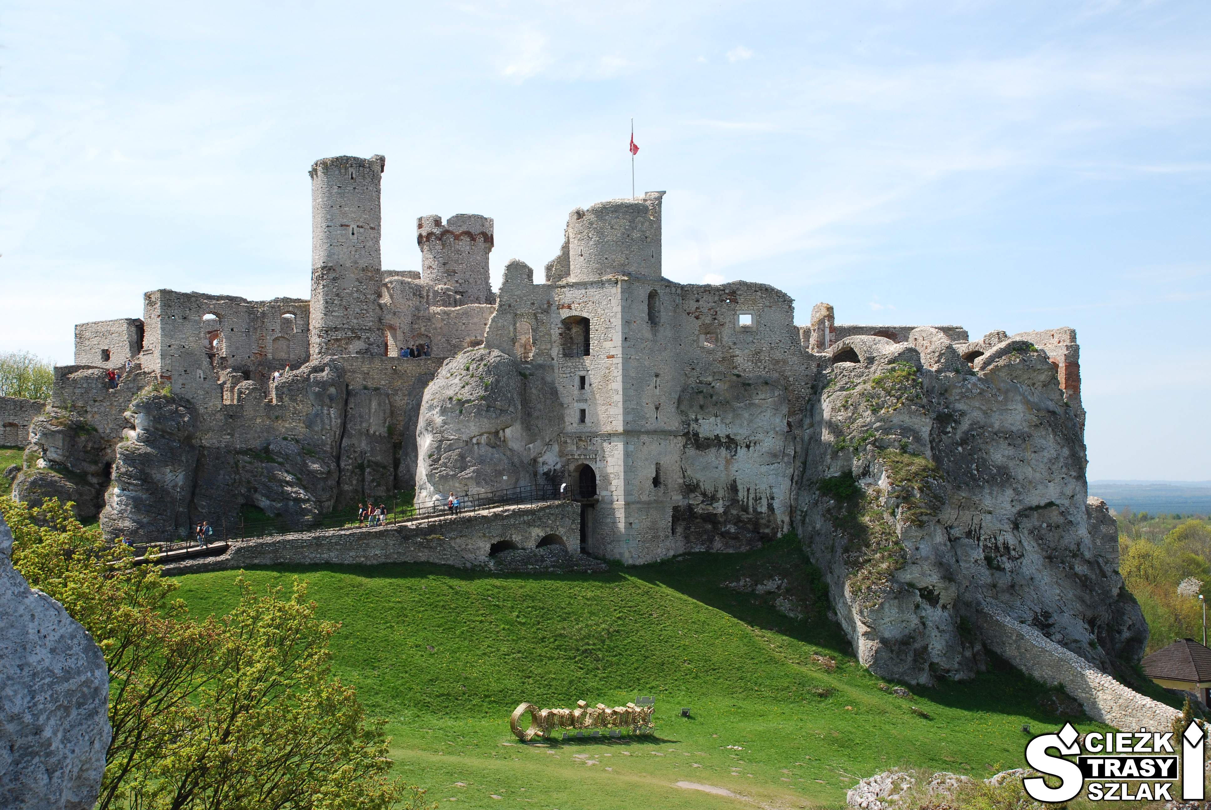 Ruiny zamku wzniesionego na skale wapiennej z dużym złotym napisem Ogrodzieniec na wzgórzu