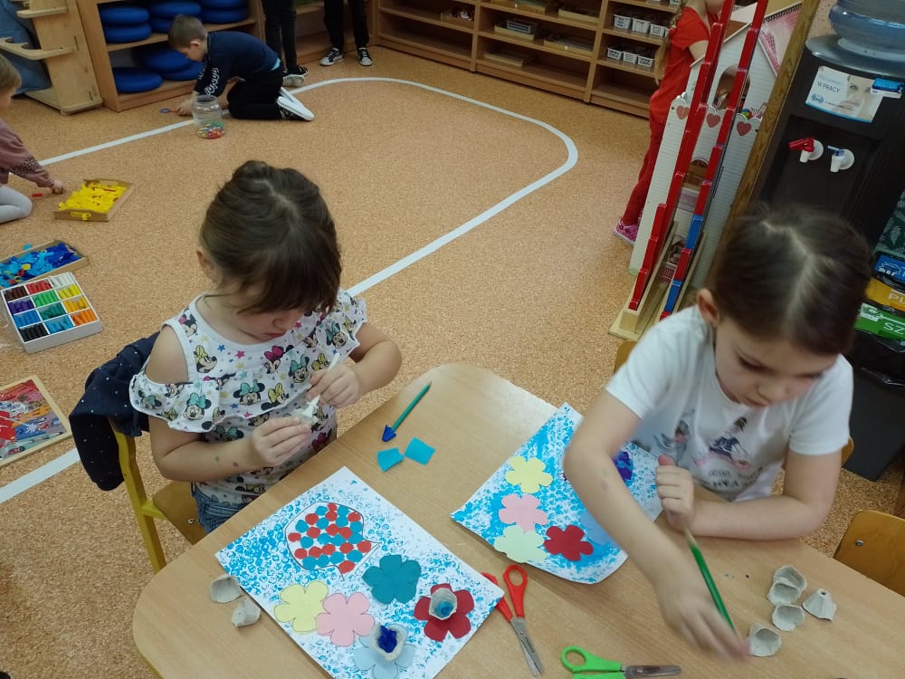 Dziewczynki naklejają kolorowe kwiatki na kartkę oraz malują farbami.