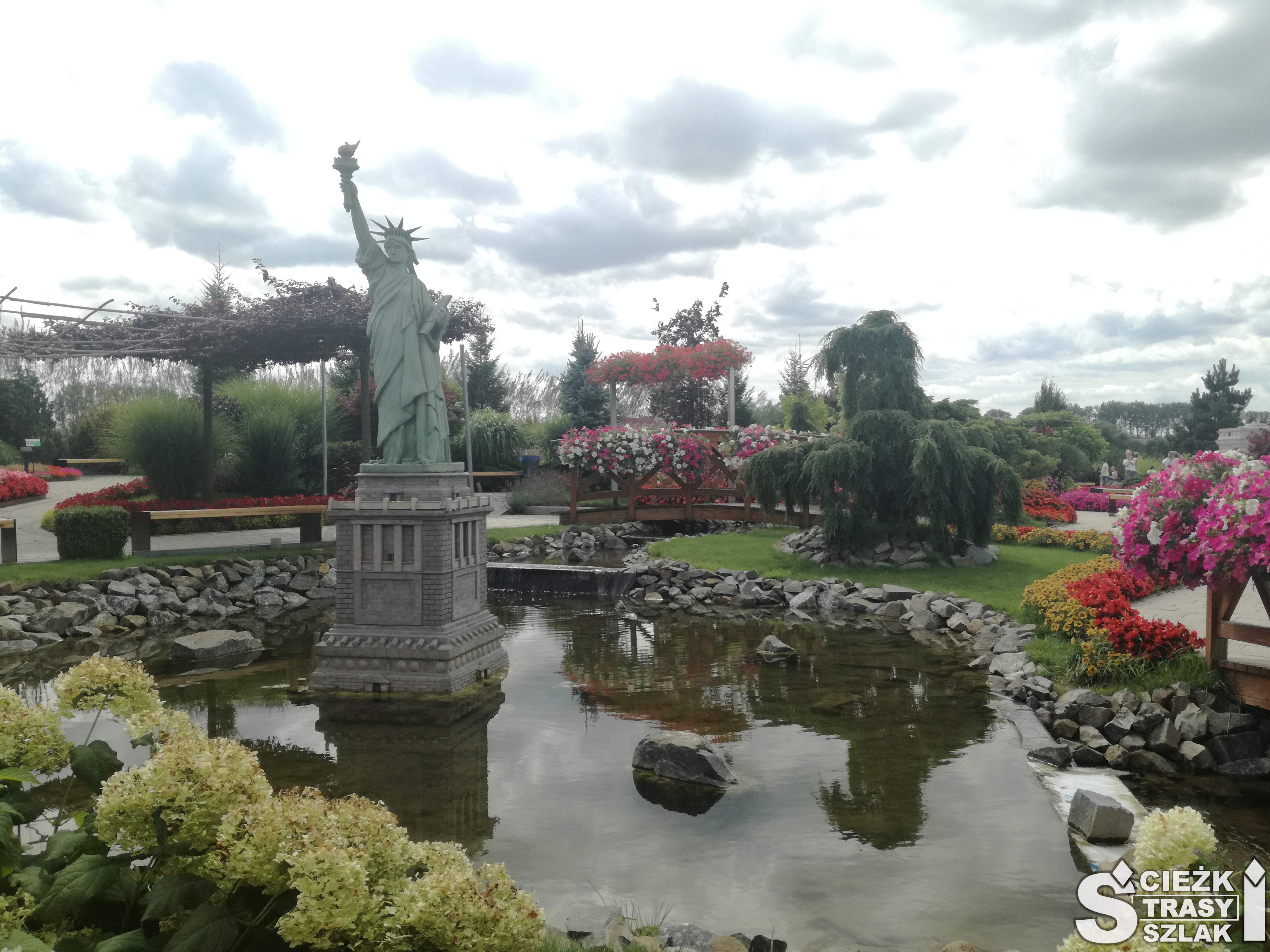 Miniatura Statuy Wolności ze Stanów Zjednoczonych otoczona stawem wyłożonym z kamienia i kolorowymi drzewami i kwiatami w Kłodzku