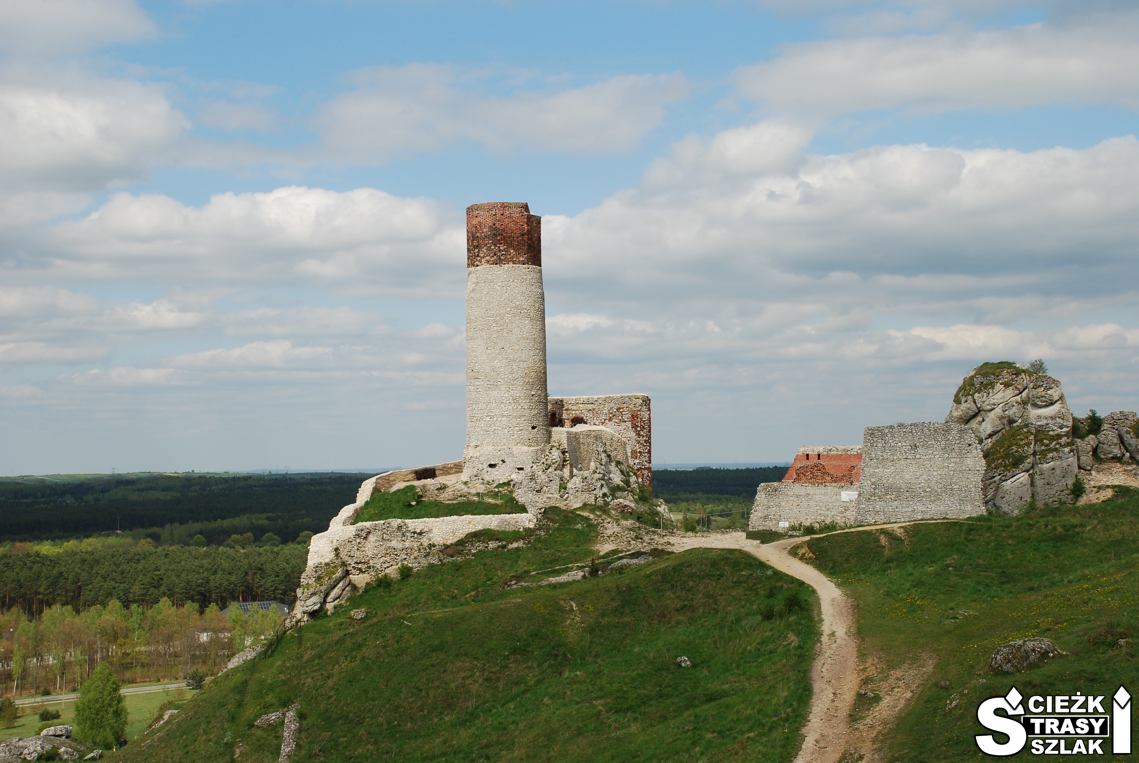 Wydeptania ściezka na zielonym wzgórzu, prowadząca do wysokiej wieży cylindrycznej przy ruinach Zamku w Olsztynie