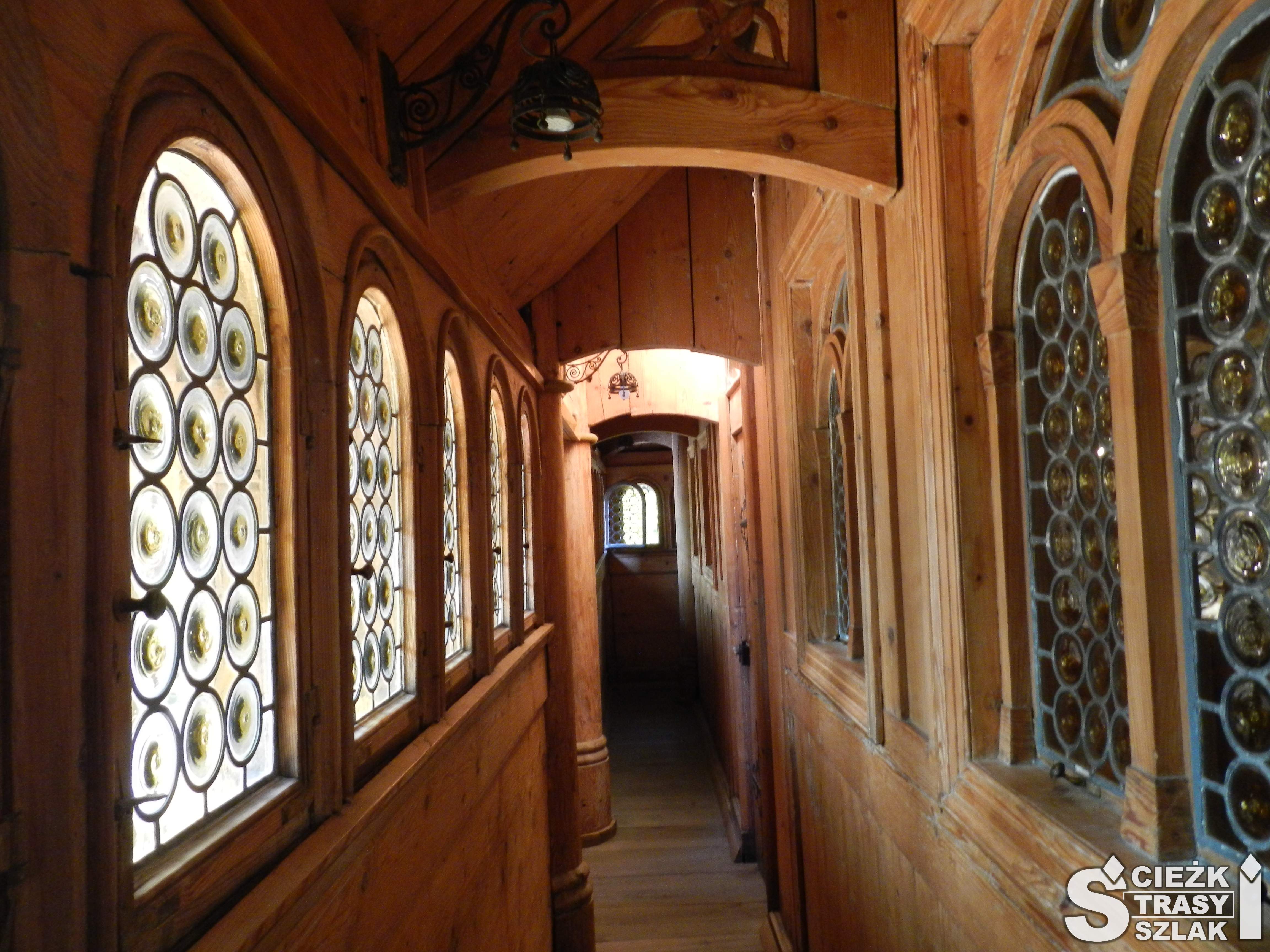 Wąski korytarz wykonany z drewna i doświetlony z zewnątrz przez zdobione witrażami okna