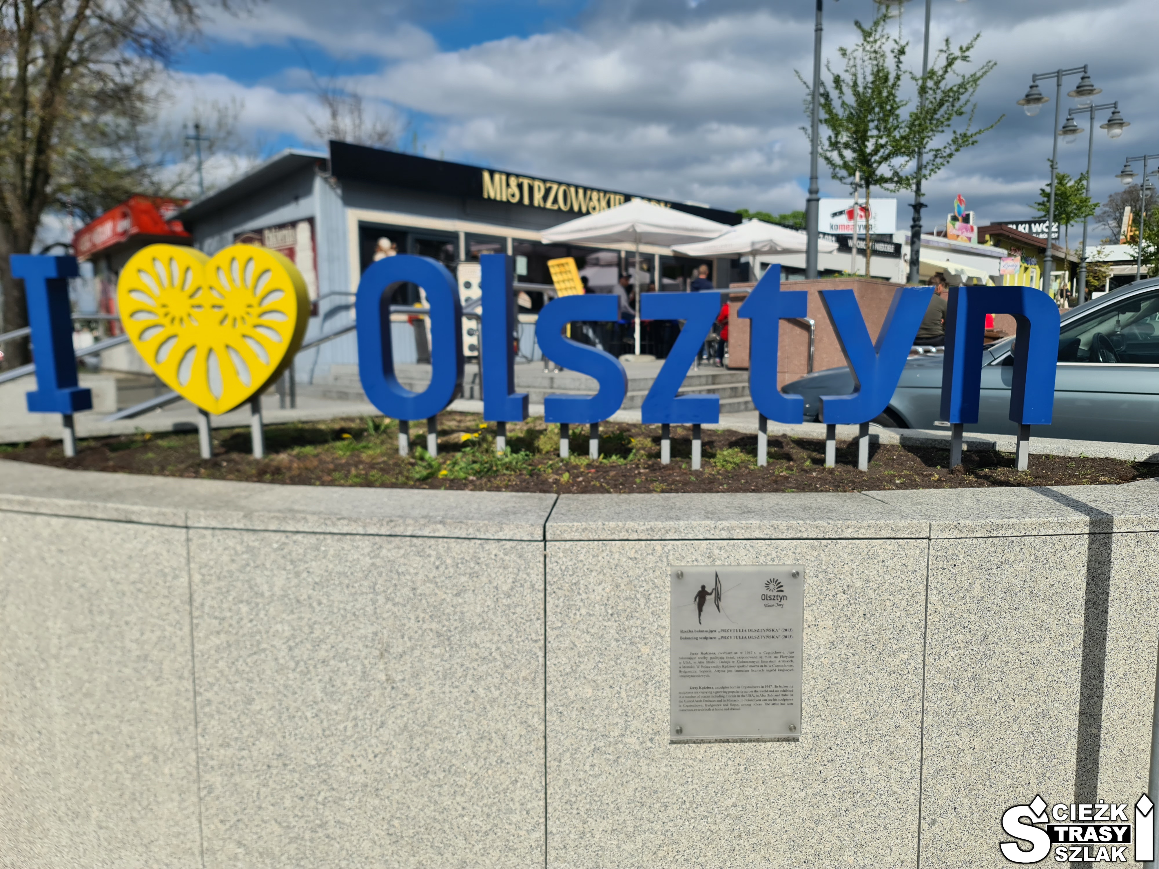 Duży, niebieski napis I love Olsztyn koło Częstochowy na rynku głównym z rzeźbą balansującą “Przytulia Olsztyńska”
