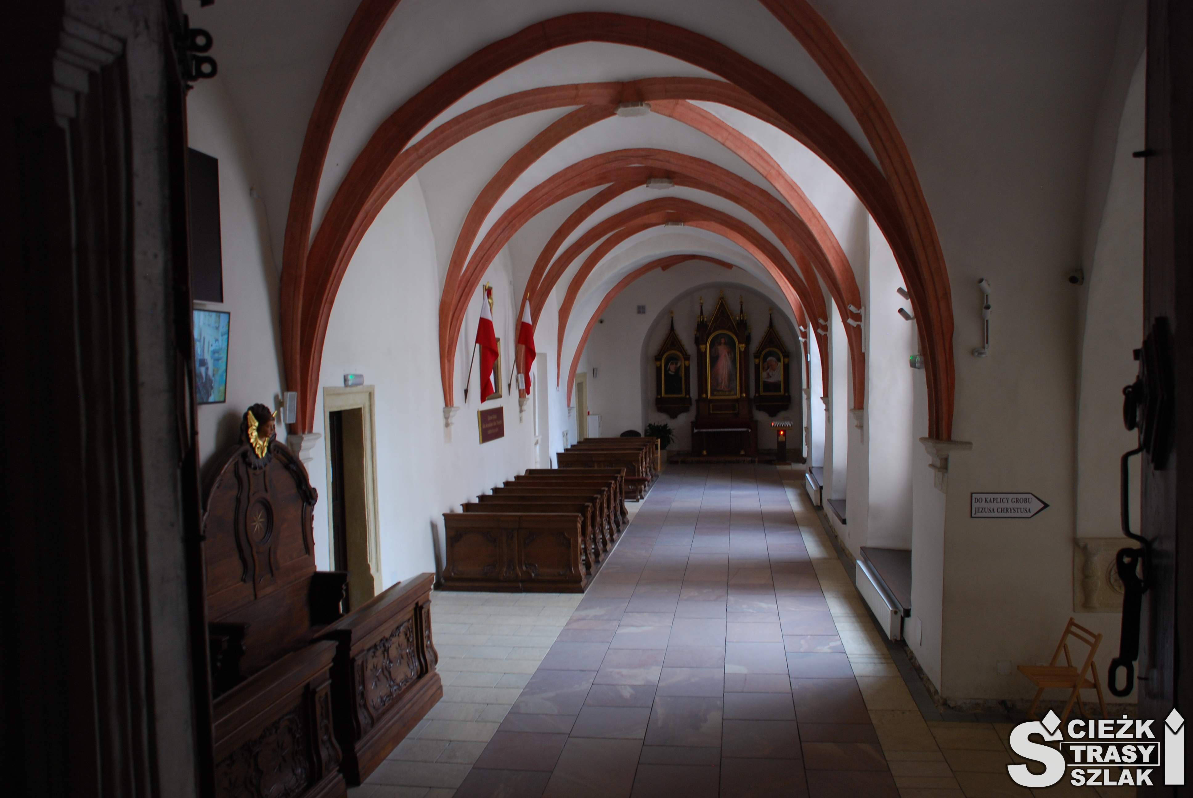 Długi korytarz z łukowatymi sklepieniami i ustawionymi przy ścianie konfesjonałami i ławkami przed ołtarzem z obrazem Jezusa Miłosiernego