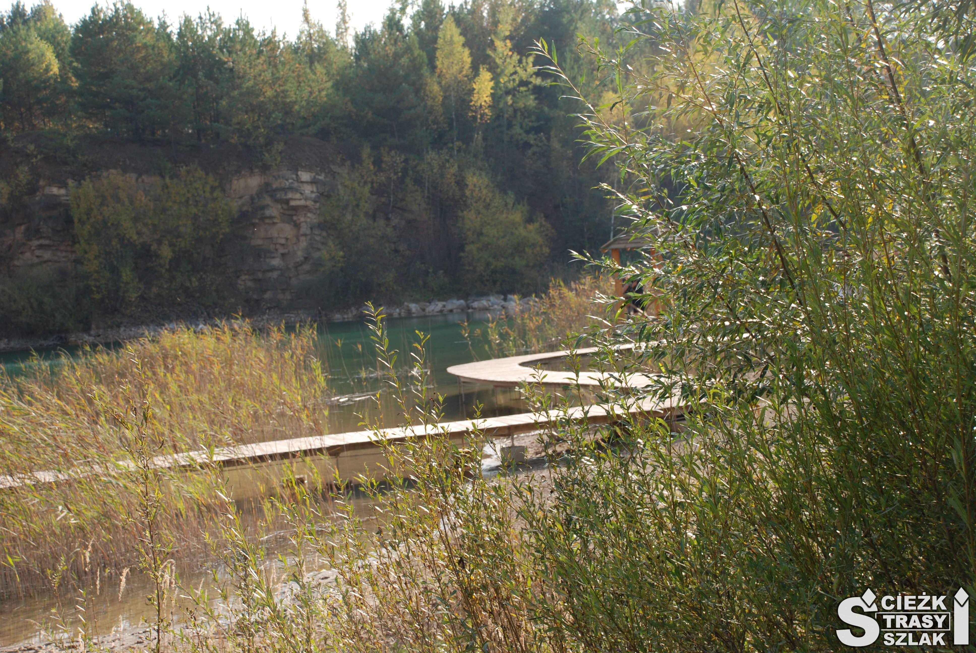 Drewniana kładka kręta nad taflą lazurowego jeziora otoczonego skarpami kamieniołomu i krzewami w Arboretum w Jaworznie