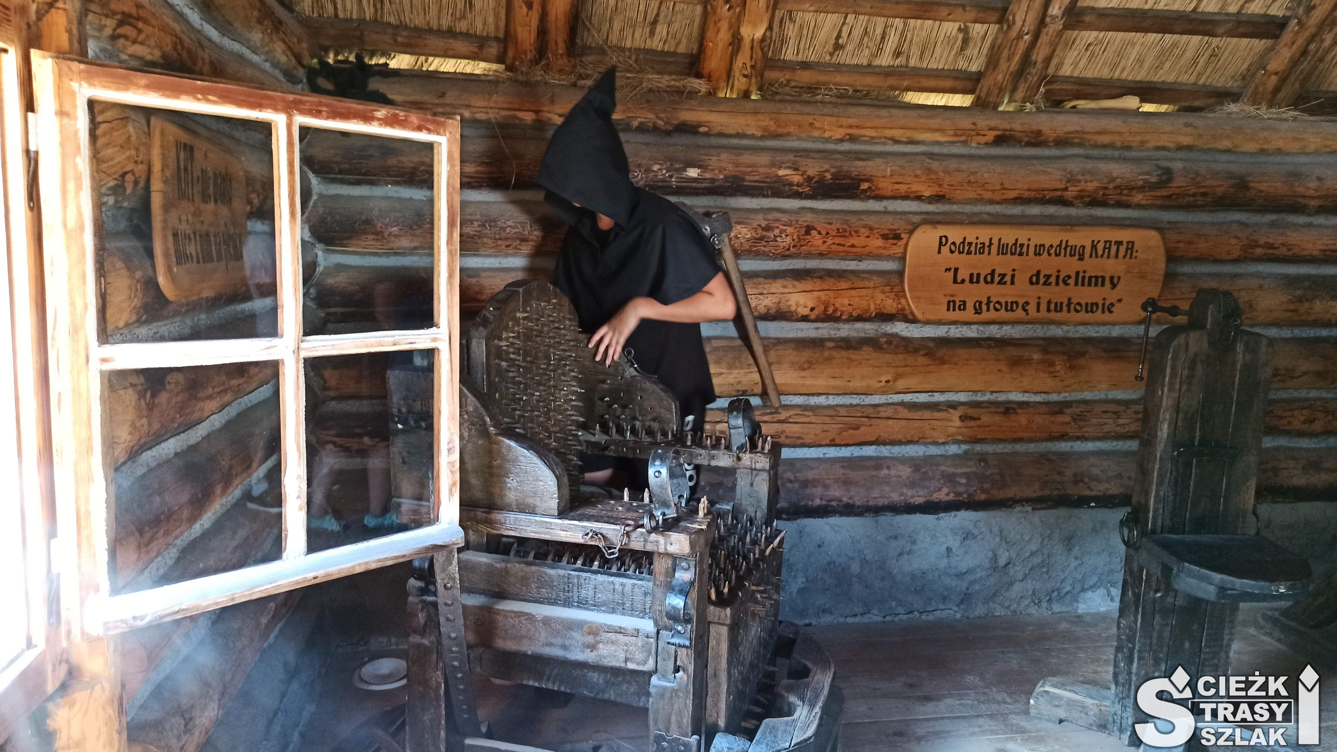 Wnętrze Chaty Kata w krzesłem do tortur, przy którym stoi gospodarz - ubrany na czarno kat
