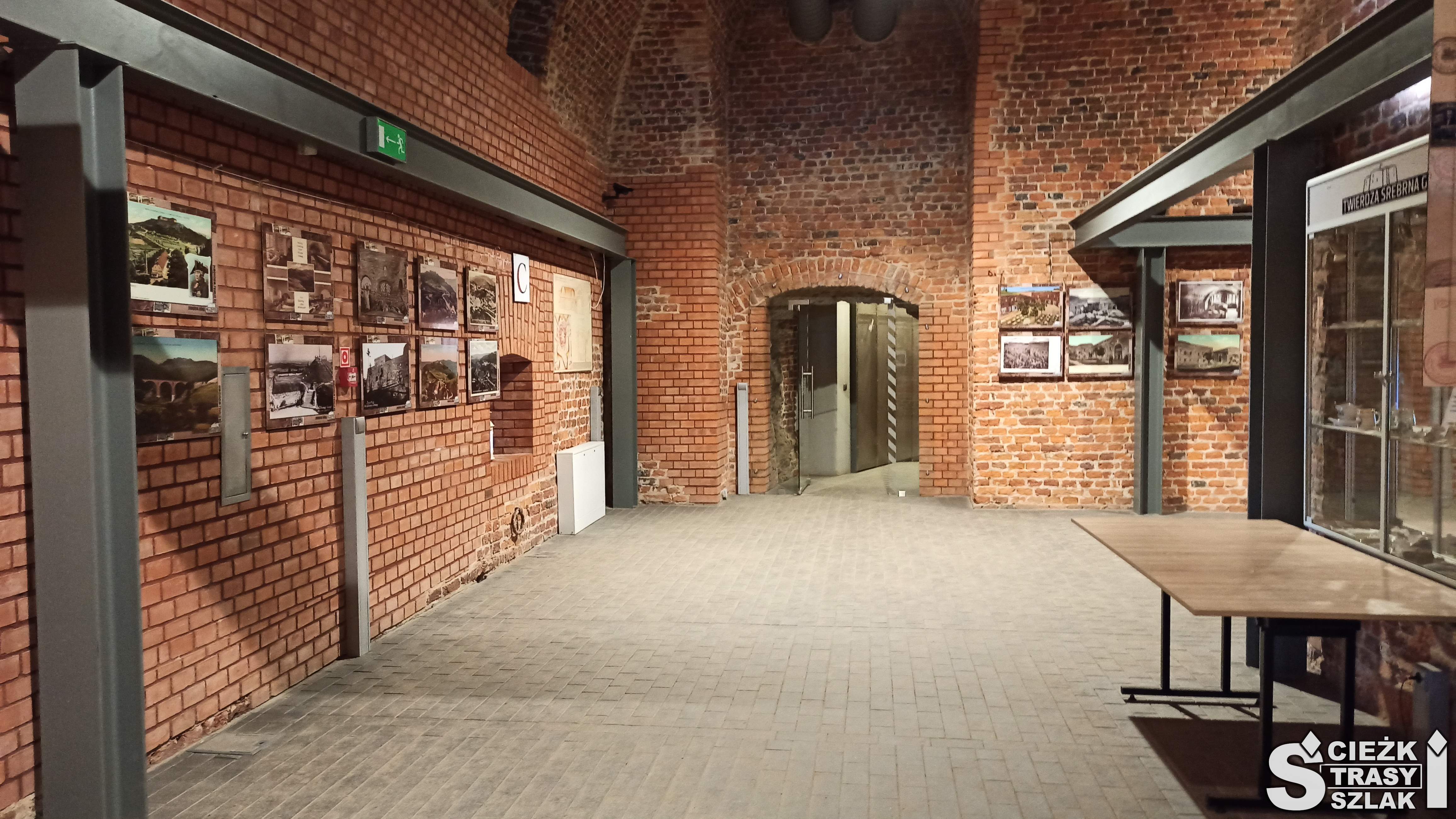 Podziemia Twierdzy Srebrna Góra z galerią zdjęć na ceglanej ścianie i szklanych regałach przed łukowatym przejściem