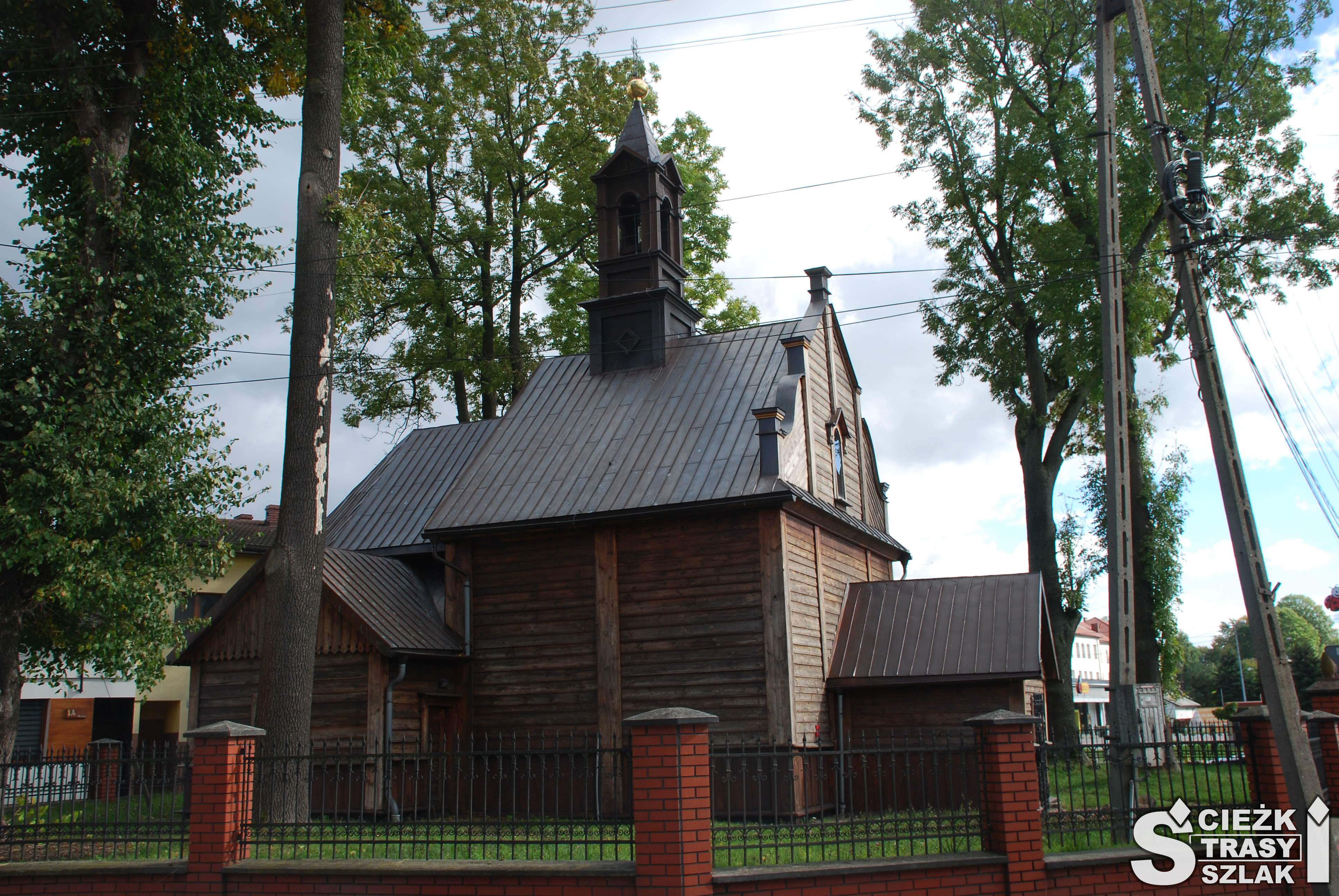 Drewniany kościół Mariacki wielkości kapliczki przydrożnej za ogrodzeniem wśród drzew na szlaku architektury drewnianej Małopolski
