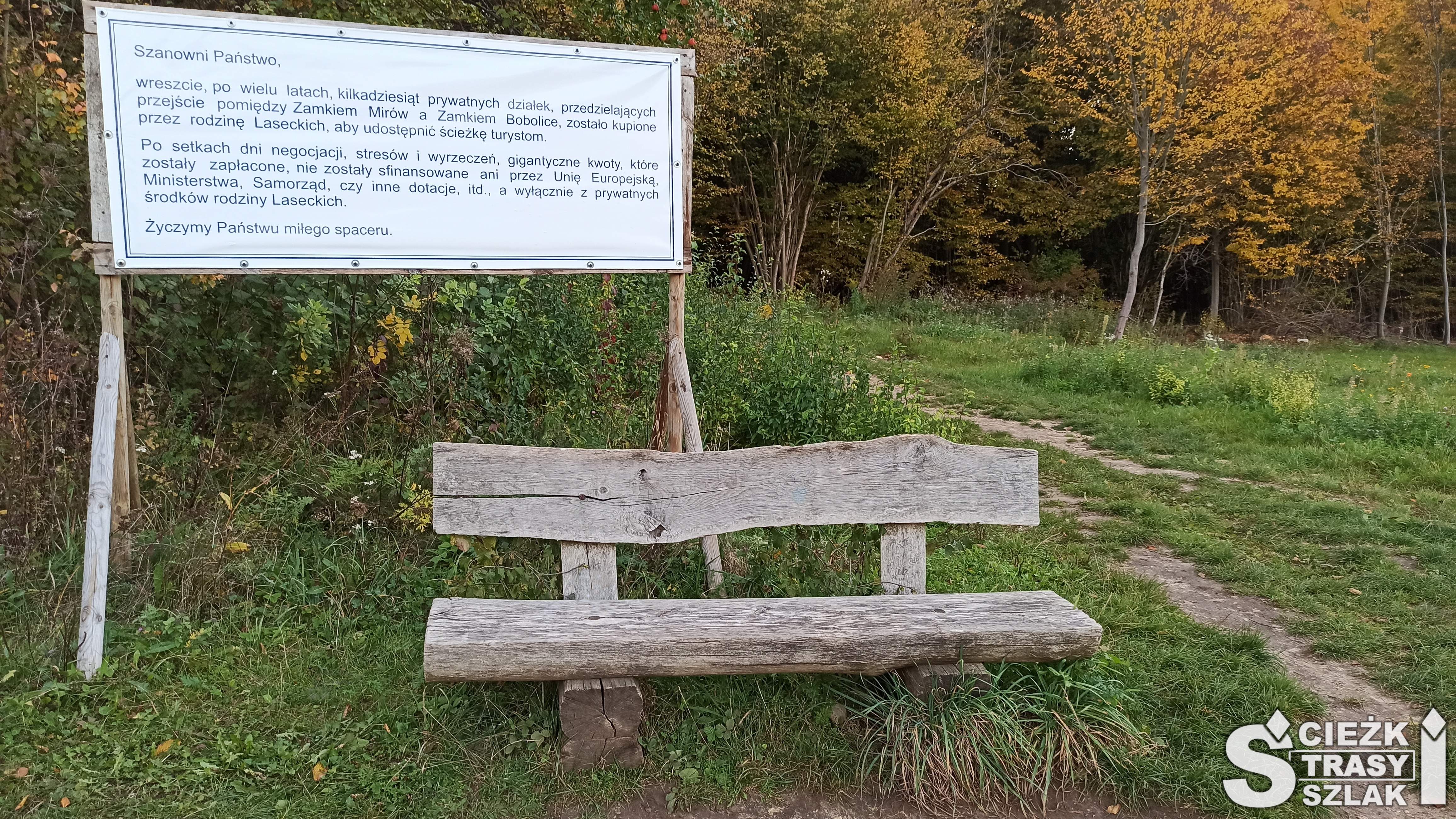 Drewniana ławka na tle tablicy informacyjnej o otwarciu czerwonego szlaku królewskiego przy ścieżce łączącej Zamek w Mirowie z Zamkiem Bobolice