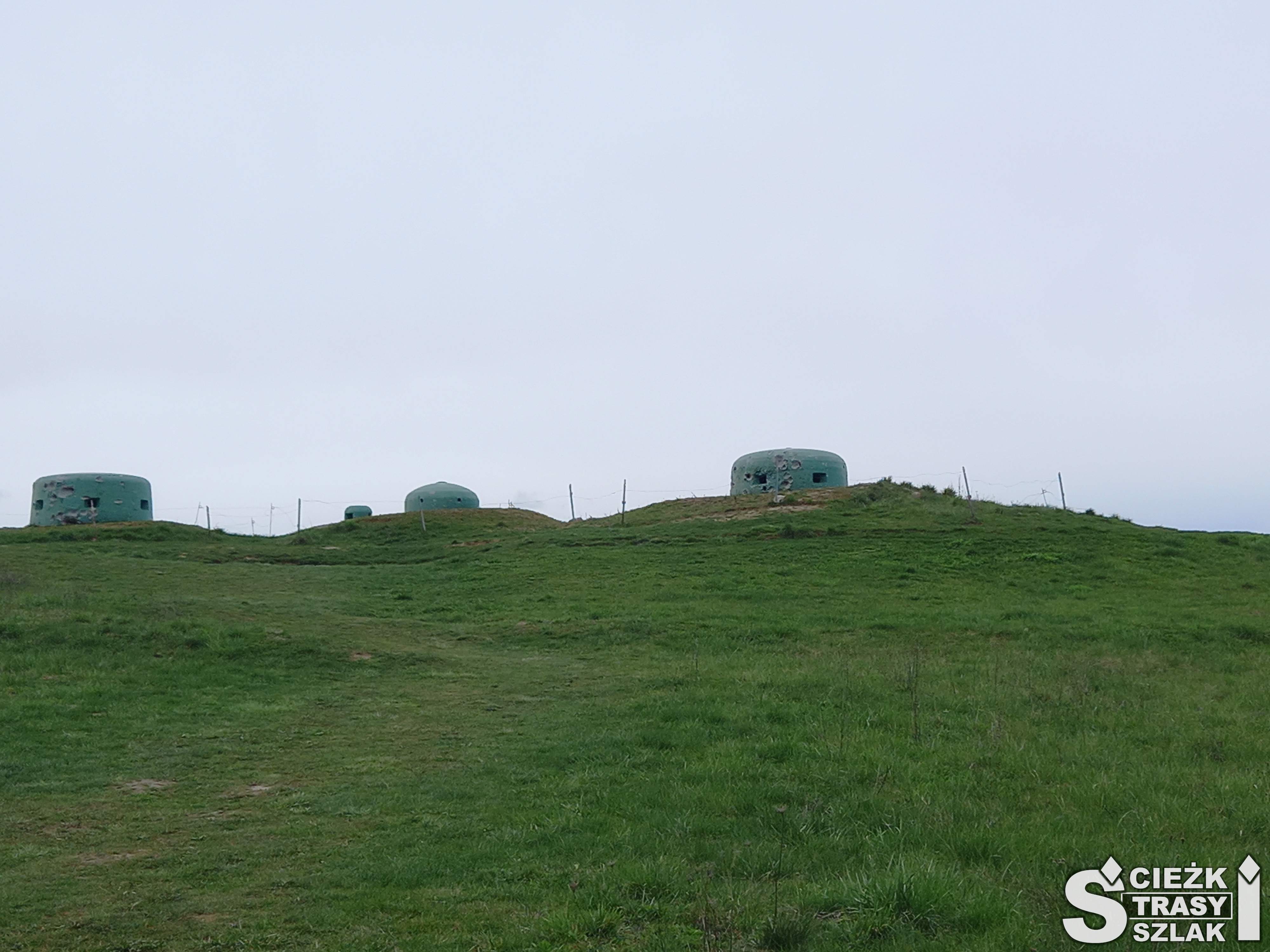 Trzy betonowe bunkry, walce z otworami na broń wystające ponad powierzchnię ziemi na zielonym wzgórzu