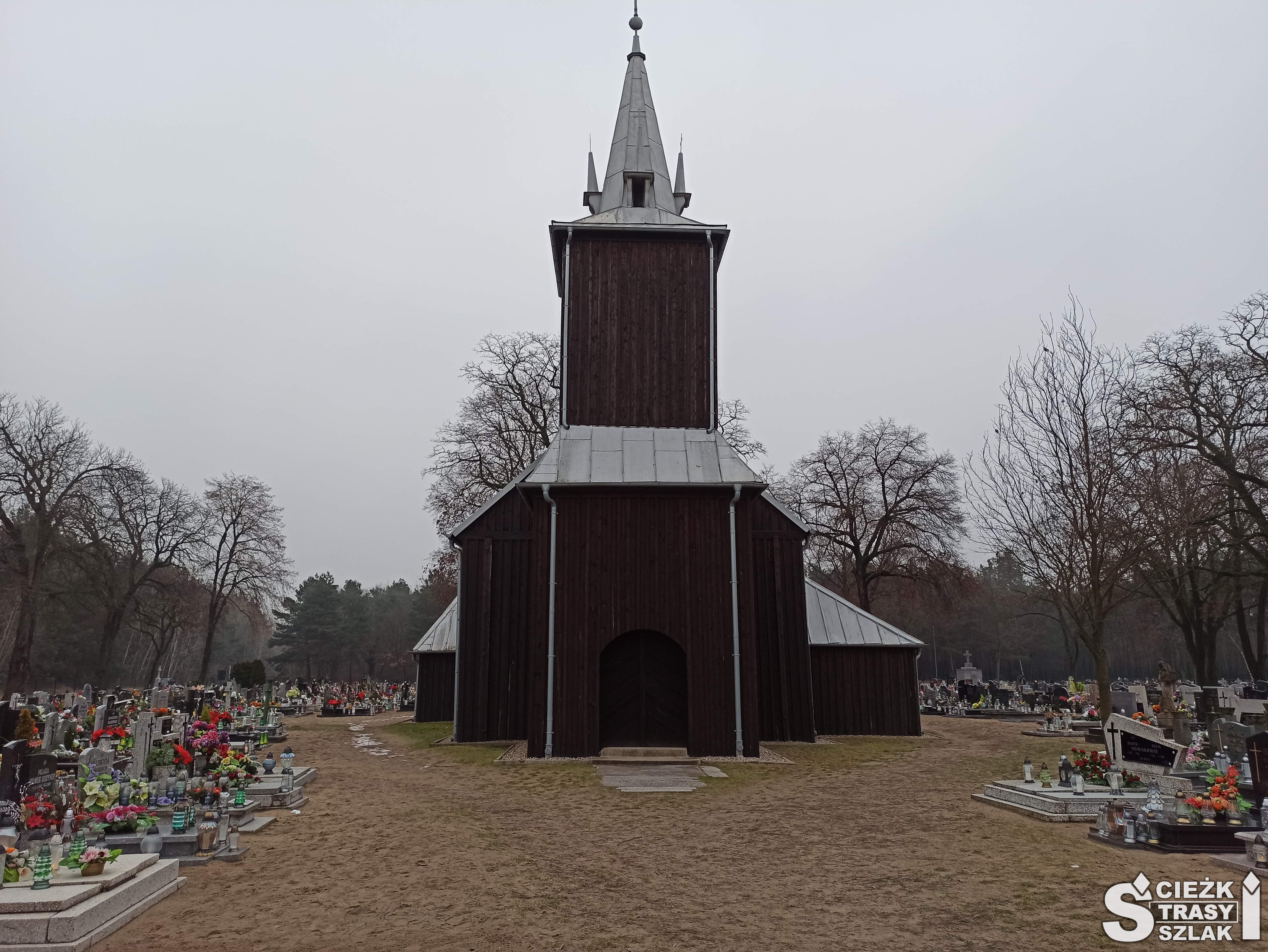 Wejście do drewnianego kościoła w Kębłowie z wysoką wieżą kościelną na cmentarzu