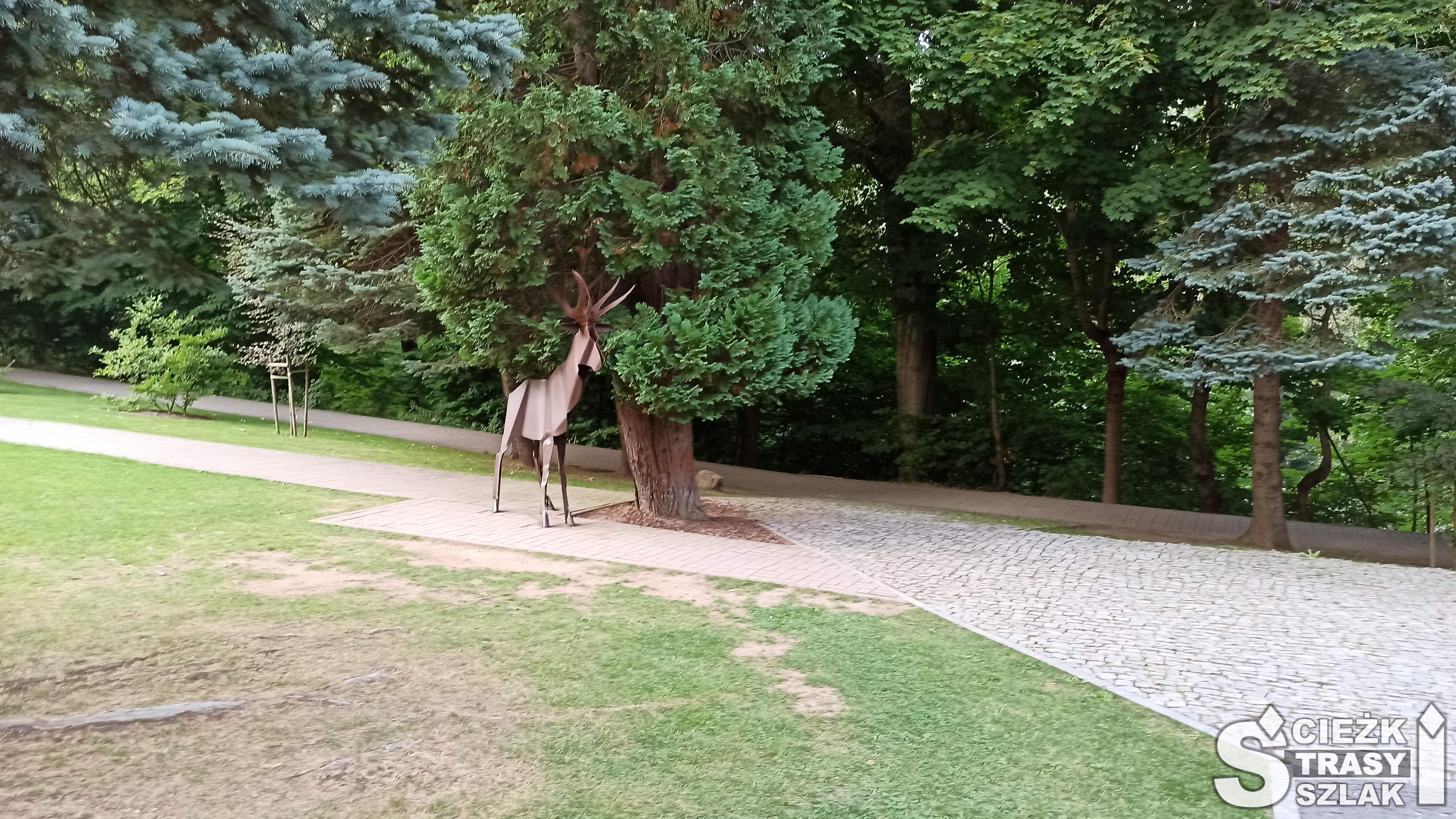 Rzeźba stalowego jelenia odpoczywającego w cieniu drzew rosnących w parku miejskim przy Zaporze na Łomnicy