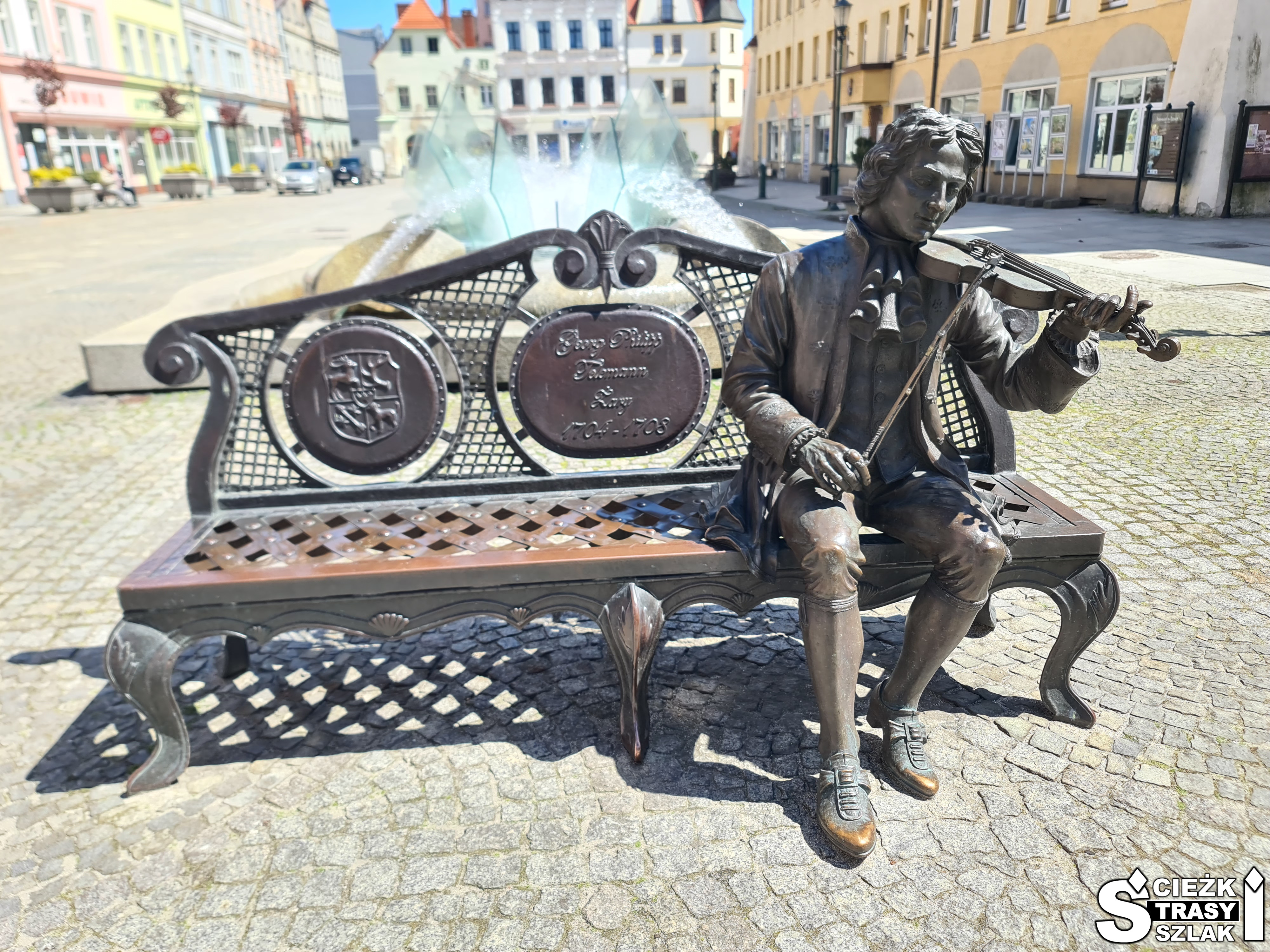 Telemann z brązu na ławeczce przy ratuszu i nowoczesnej fontannie w Żarach trzymający skrzypce