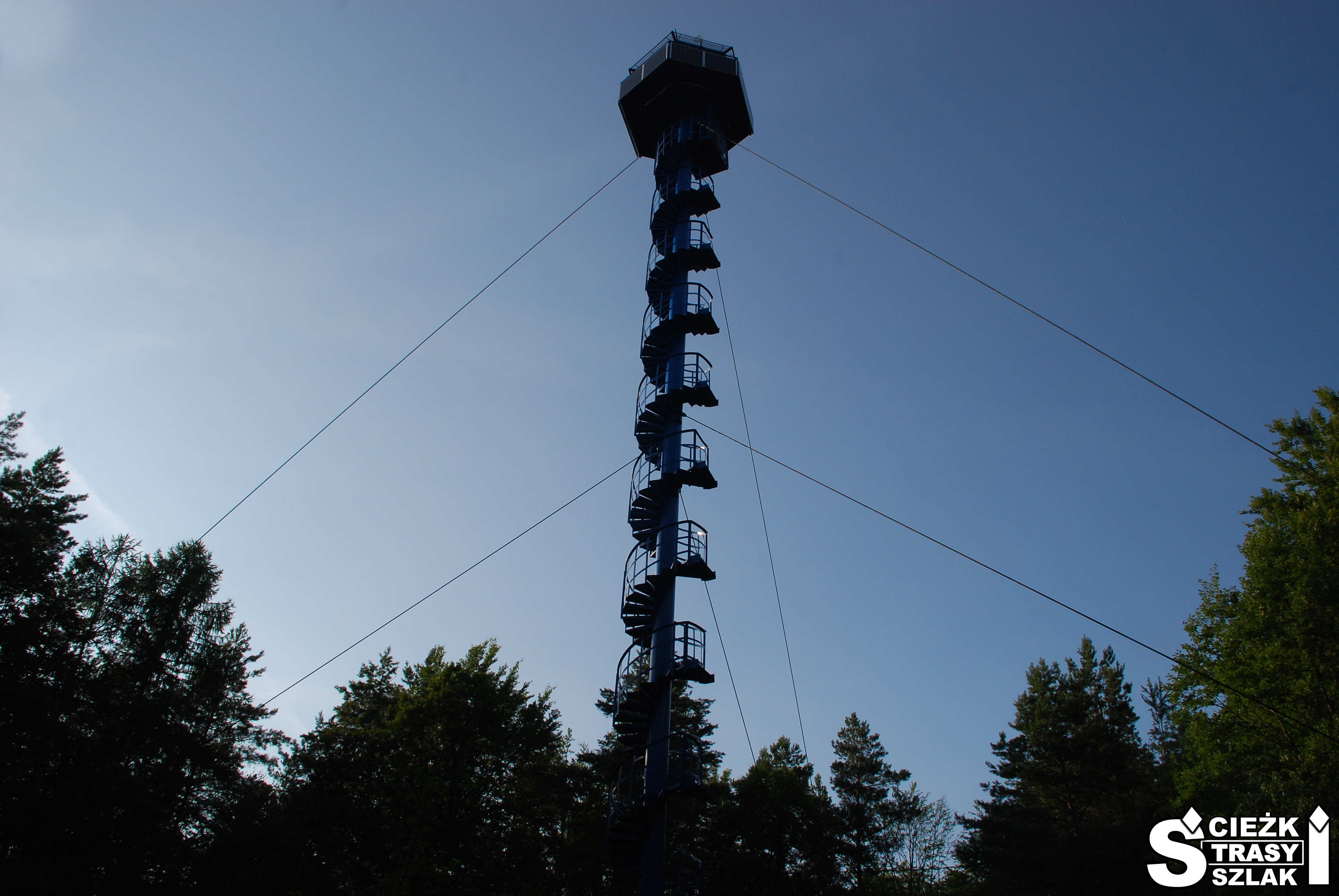 Wysoka wieża P-POŻ z krętymi schodami przypominająca gwint śruby na tle drzew Zielonego Lasu w Żarach