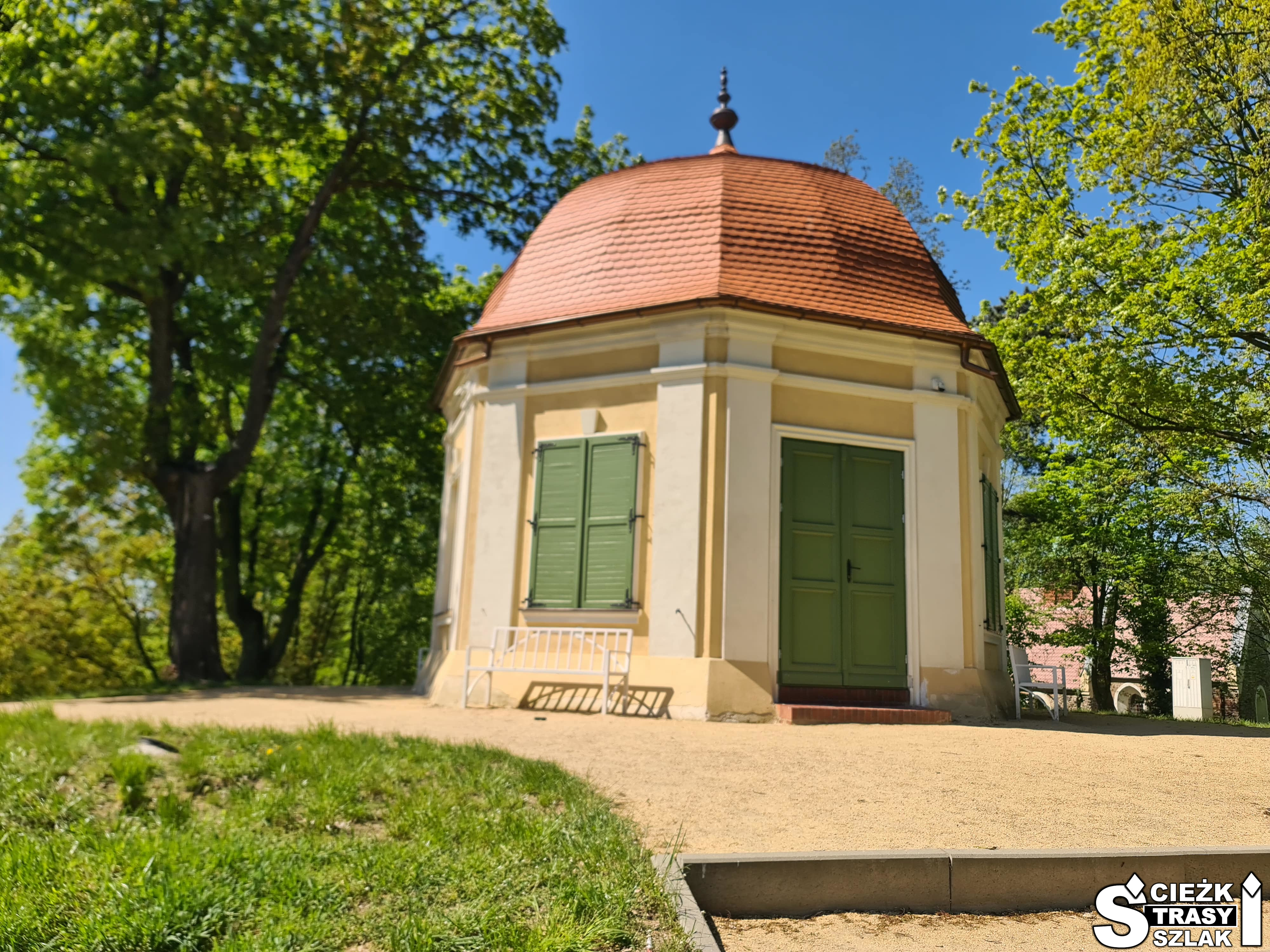 Mały domek na planie sześciokąta z zielonymi okiennicami i drzwiami w zespole zamkowo-pałacowym w Żarach - Domek Winny