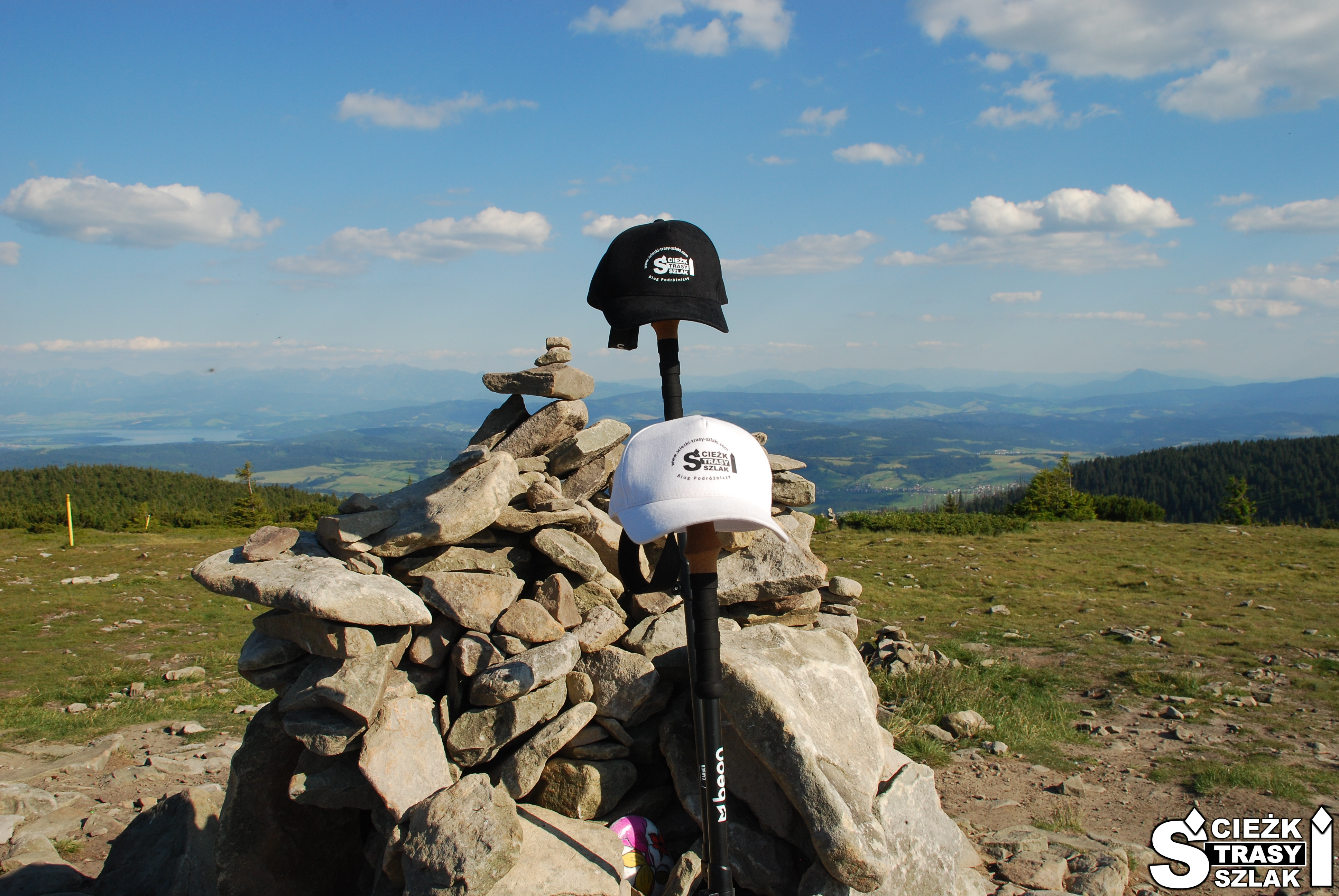 Czapeczki z logo bloga podróżniczego na kijkach trekkingowych opartych o kopiec z kamieni na szczycie Góry Pilsko