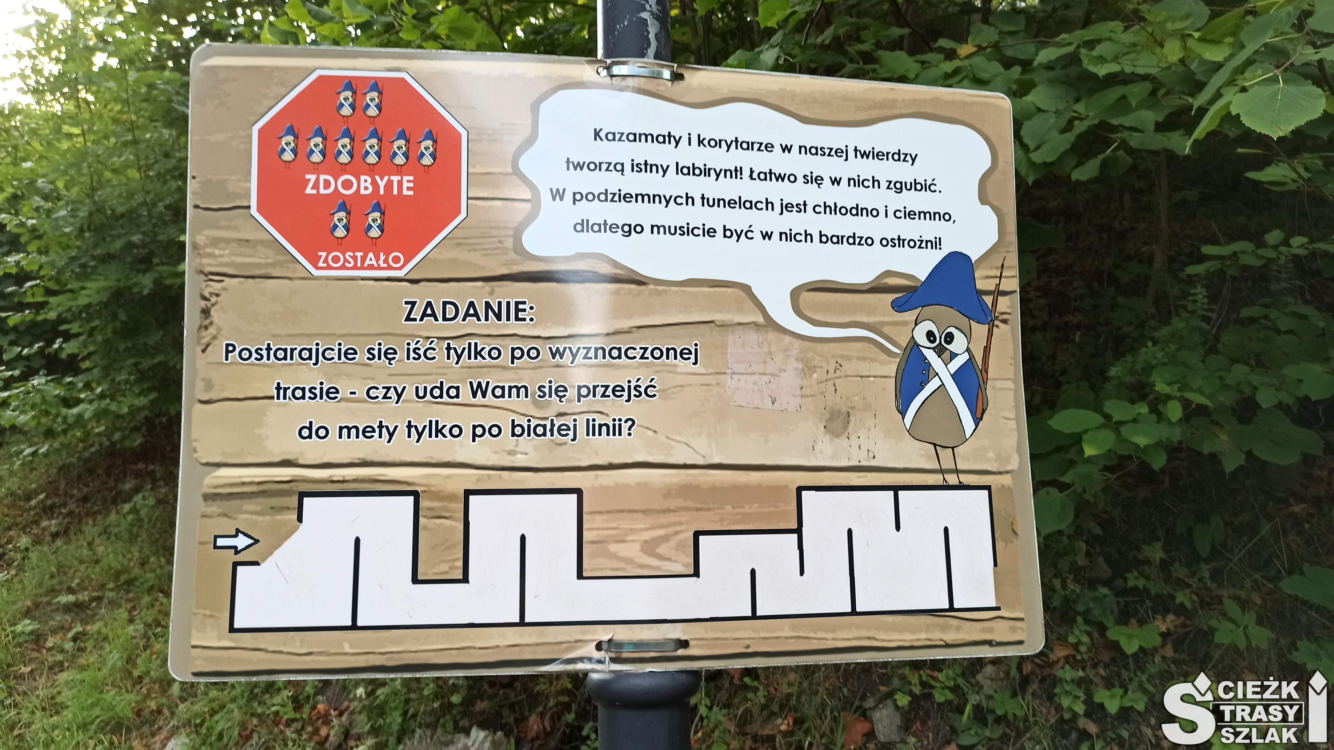 Tablica informacyjna na ścieżce dydaktycznej z rebusem dotyczącym labiryntu wymalowanego białą kredą na ulicy Krętej w Srebrnej Górze