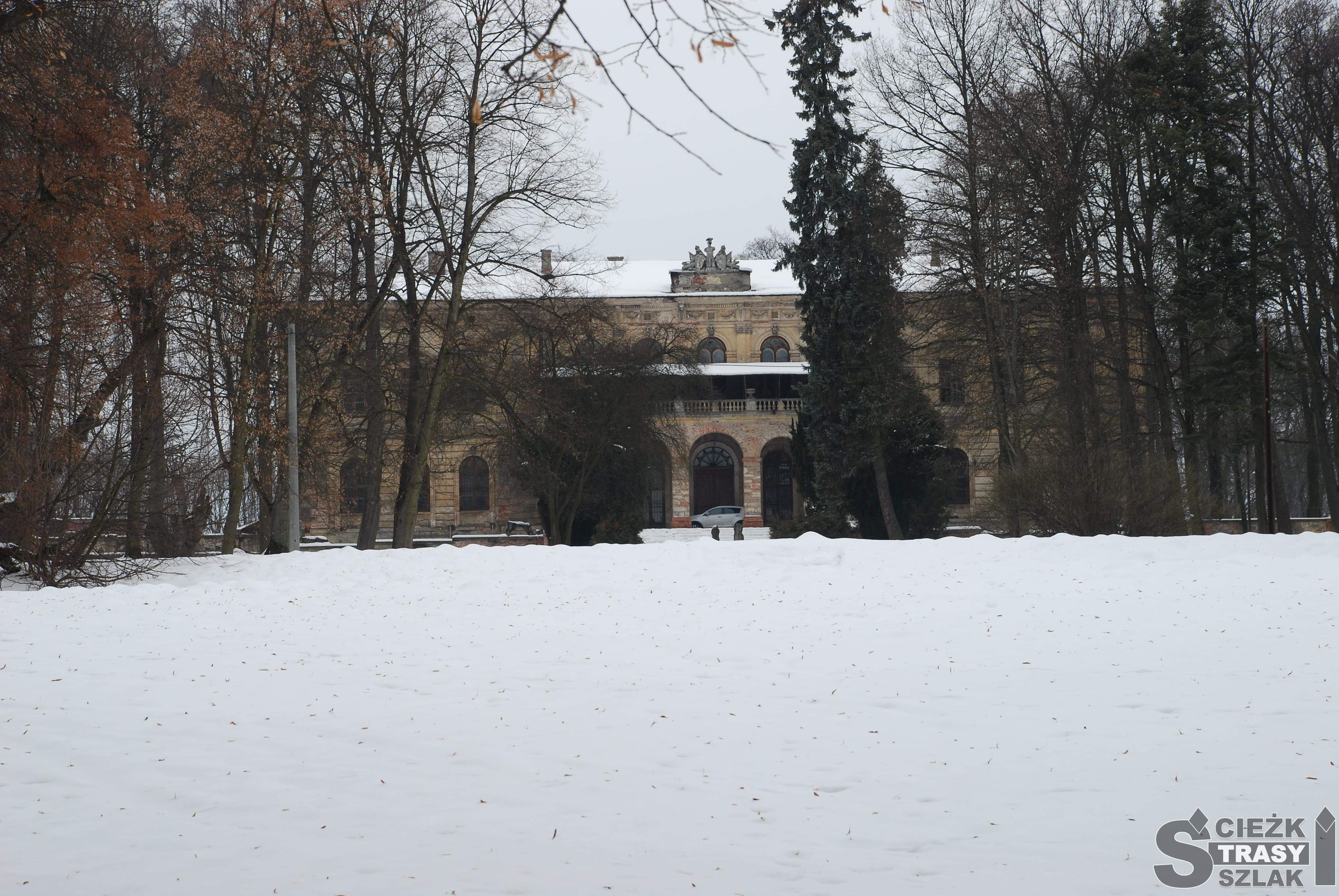 Pałac w Pilicy z wejściem z łukowatym kolumn otoczony drzewami w parku pokrytym śniegiem